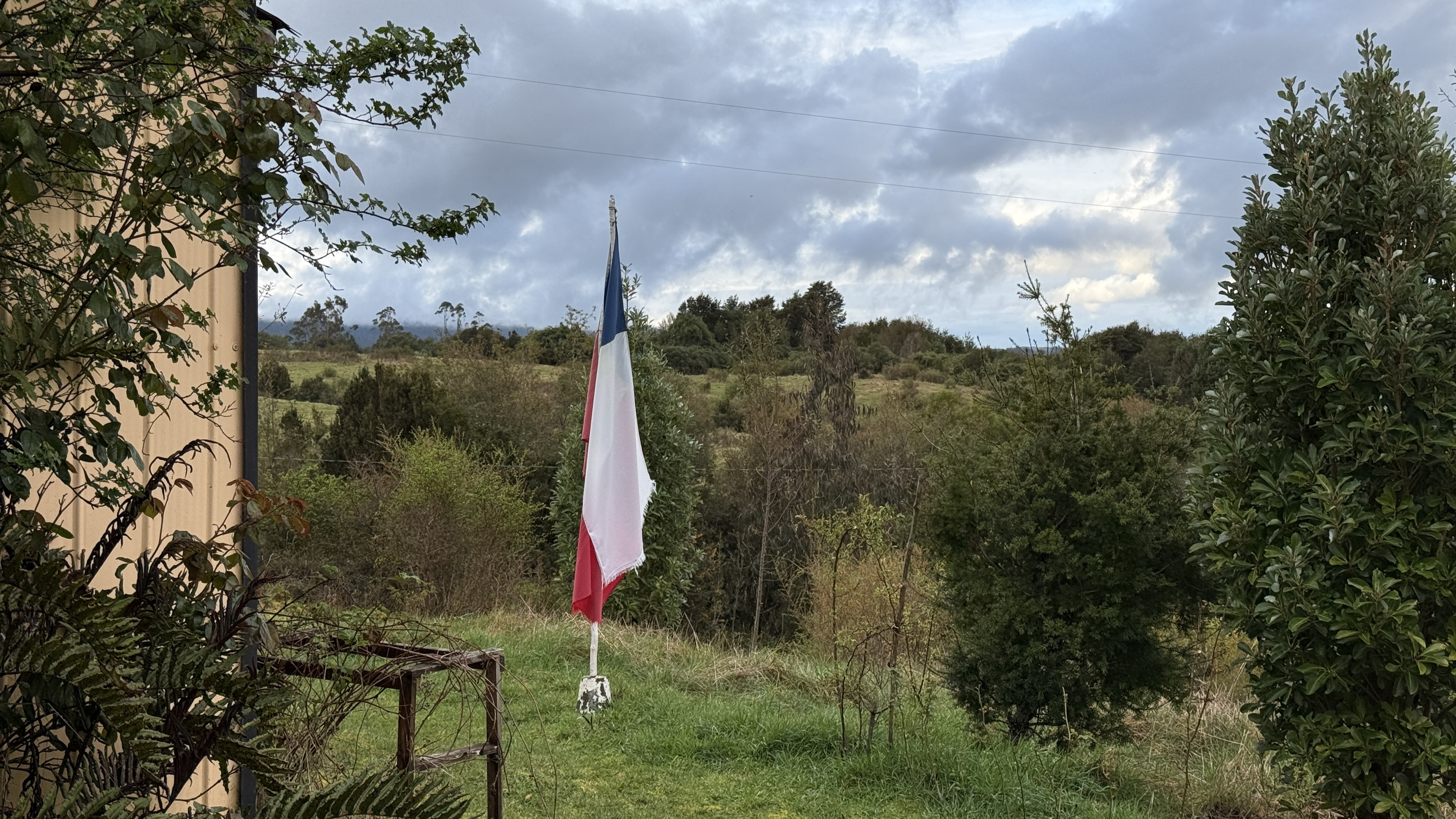A weathered Chilean flag on a fence post in a green rural clearing under a cloudy sky.