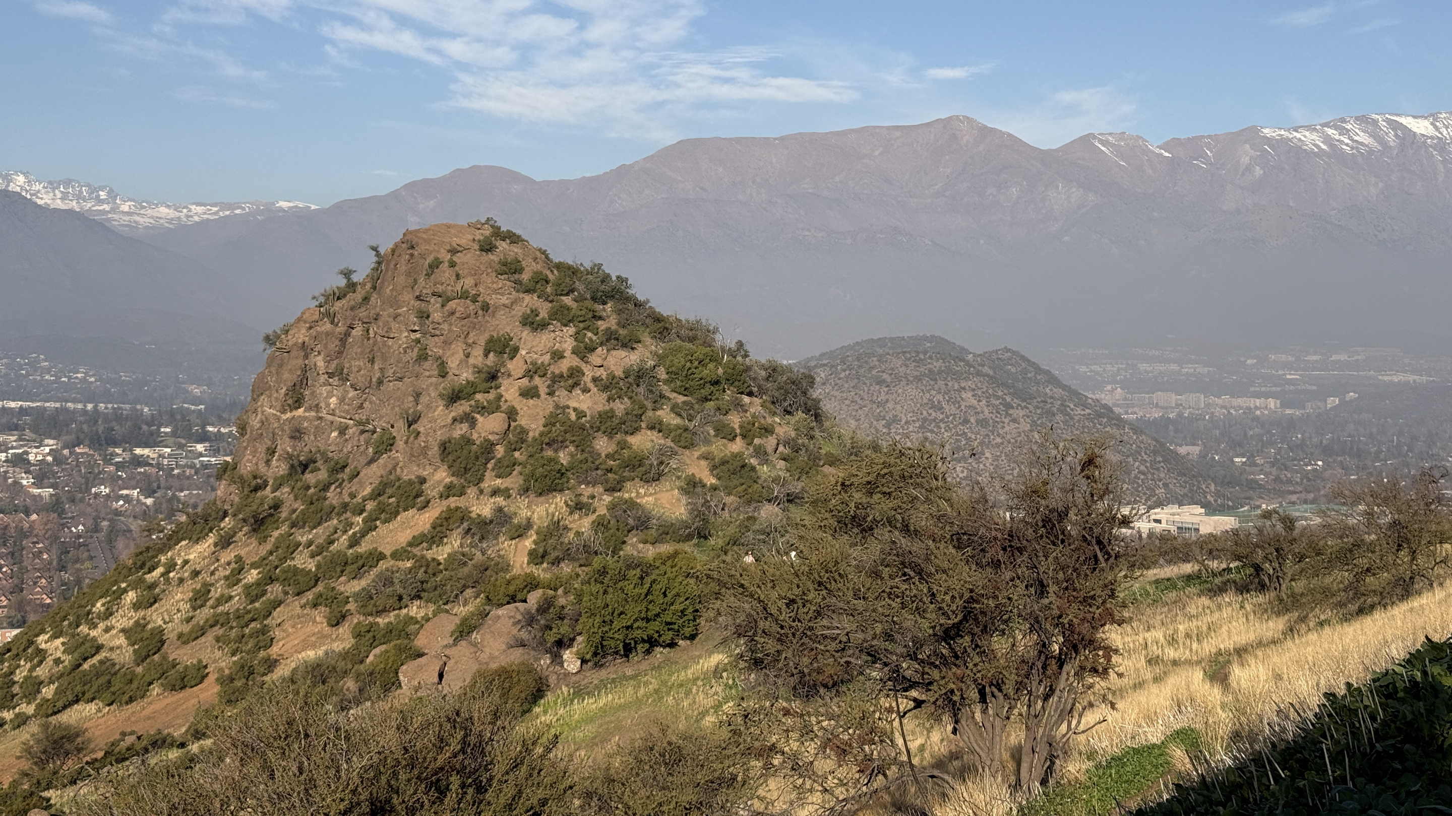 A rocky summit over Santiago's eastern suburbs through the haze, with the Andes behind.