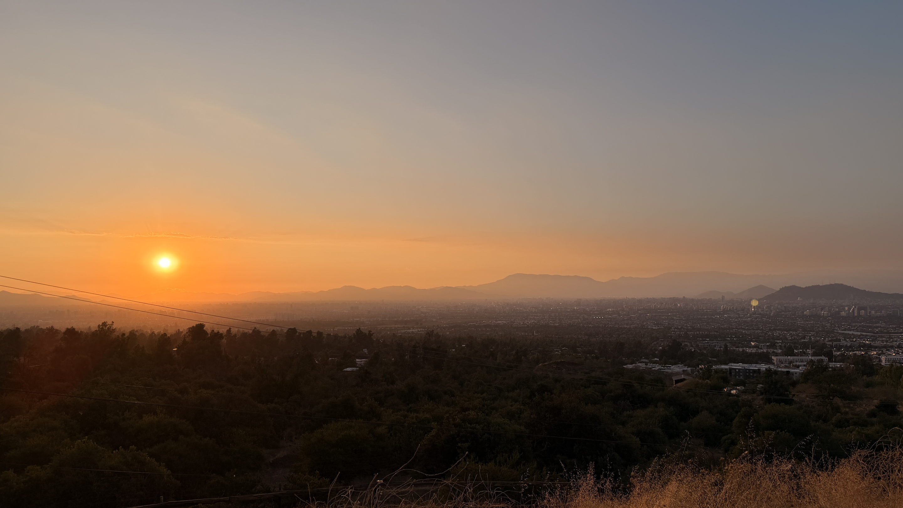 The sun setting into haze over the Santiago basin, seen from a forested hillside.